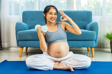 Young pregnant Asian woman about to become a mother, wearing a sports bra, exercising, and sitting lifting dumbbells up and down slowly to flex her arm muscles before giving birth on a mat, Looks OK