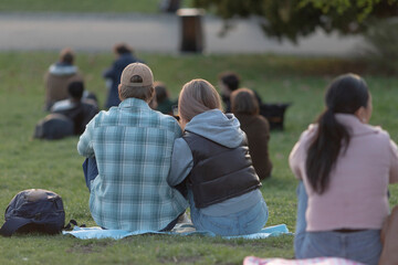 A couple sits closely together on a cozy blanket in a park, surrounded by other people who are also...