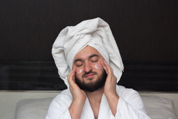 Male portrait. Young adult man with closed eyes beard in robe, white towel on head applies milk...