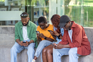 Black friends sit on bench, glued to smartphones, chatting and scrolling through social media, ignore each other. Gadget addiction, immersed in social networks world, disconnected from surroundings 
