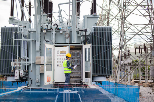 Engineer performs inspection on large transformer at electrical substation wearing safety gear and yellow helmet ensuring equipment operates safely and efficiently