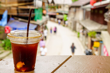 Iced coconut americano coffee on wooden table with blurry Mae Kampong street background, small village with traditional houses and beautiful nature. Destination of traveler. Chiang Mai, Thailand.