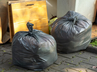 Black garbage bags on pavement next to old furniture. Waste disposal scene in urban environment,...