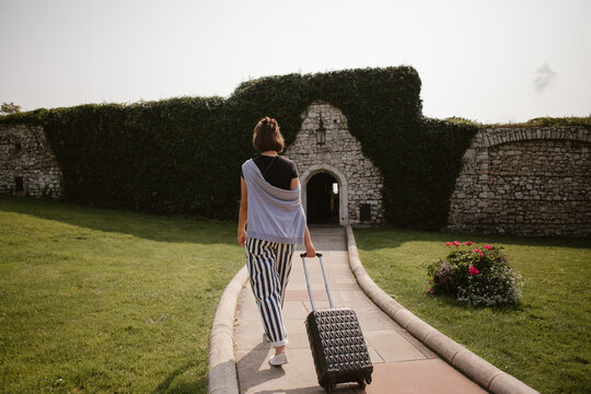 Young woman walking towards ancient building pulling trolley suitcase