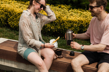 A couple enjoys a light picnic with traditional khachapuri, tea, and juice near the Bridge of Peace in Tbilisi on a sunny summer day