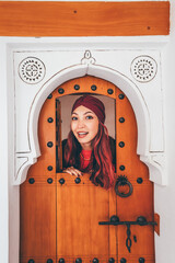 Young woman wearing a vibrant turban, gazing through an ornate wooden door, immersing herself in the rich culture of Morocco
