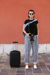 Young woman tourist holding camera with luggage posing in front of red wall