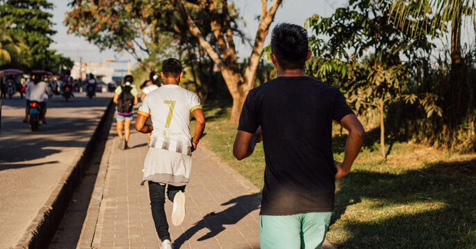 Dos j&oacute;venes corriendo por una acera junto a una pista, en un entorno urbano, con &aacute;rboles y vegetaci&oacute;n a los lados de la acera, al atardecer.