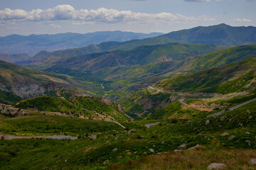 Fototapeta premium Scenic view of lush green hills and dramatic red cliffs under a bright blue sky with white clouds, showcasing the beauty of untouched mountainous nature and diverse rocky terrain.