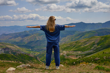 Naklejka premium Woman standing on a mountain peak with arms outstretched, enjoying a breathtaking panoramic view of green hills and valleys under a partly cloudy sky, symbolizing freedom and connection with nature.
