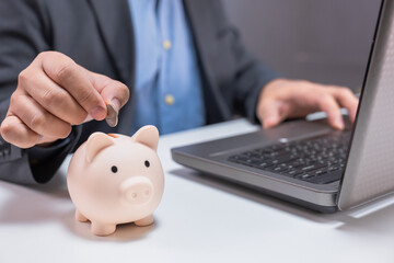 A person in business attire places a coin into a piggy bank beside a laptop, symbolizing saving...