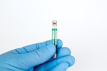 Gloved Hand Holding Medical Vaccine Vial on White Background, Dental Anesthesia Syringe.Close-up of a blue-gloved hand holding a small glass vaccine vial with a green label