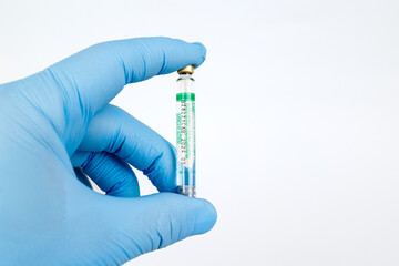 Gloved Hand Holding Medical Vaccine Vial on White Background, Dental Anesthesia Syringe.Close-up of a blue-gloved hand holding a small glass vaccine vial with a green label