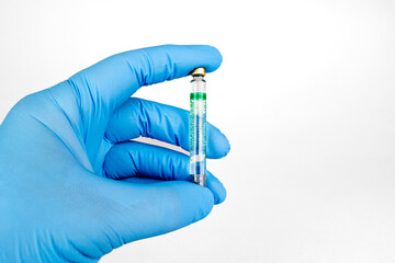 Gloved Hand Holding Medical Vaccine Vial on White Background, Dental Anesthesia Syringe.Close-up of a blue-gloved hand holding a small glass vaccine vial with a green label