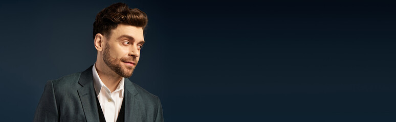 Young bearded man in dapper suit posing on dark backdrop.