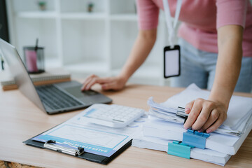 Woman holding document file stack of business document paper looking at laptop computer flicking through clients files on table, searching for and picking a file in home office.