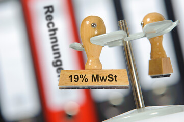 Ein Holzstempel ist beschriftet mit dem Aufdruck 19% MWST vor einem Regal mit vielen Aktenordnern