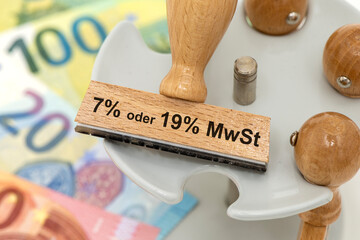 Ein Holzstempel ist beschriftet mit dem Aufdruck 7% oder 19% MWST über vielen Euro Banknoten