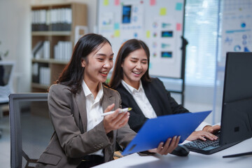 Two Asian businesswomen are working together in the office, using a laptop and documents, discussing work and smiling