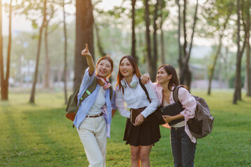 Three happy female students with backpacks and books walking in a park, one pointing at something...