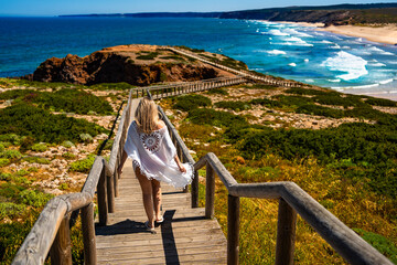Visiting Algarve coast in Portugal. Beautiful middle-aged woman tourist walking down wooden stairs to sandy Bordeira beach and enjoying vacations over ocean on summer day. Back view	