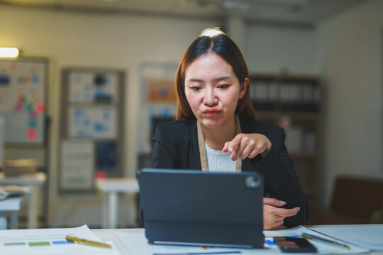 Asian businesswoman in suit having video conference and pointing at tablet sitting at desk in office