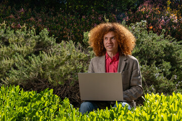 Young man with curly hair working on a laptop outdoors, surrounded by lush greenery. Concept of sustainable remote work, eco-friendly business, and digital nomad lifestyle