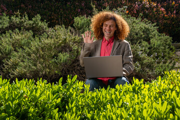 A young redhead executive dressed casually with afro hair waving on a video meeting. He is working on his laptop in the middle of a public park surrounded by plants. Sustainability and technology