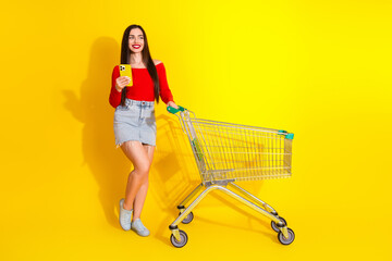 Young woman shopping with cart holding smartphone against yellow background expressing joy and...