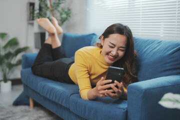 Happy asian woman lying on the couch using smartphone, browsing internet, watching video, chatting...