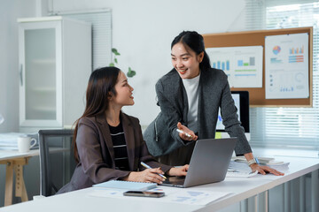 Two businesswomen are discussing work using laptop computer and documents with charts and graphs showing the performance of their company