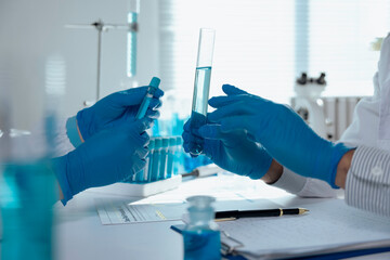 Two scientists wearing blue gloves are comparing test tubes containing blue liquid, conducting...