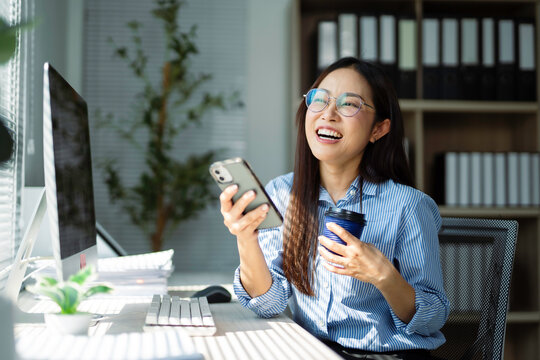 Happy businesswoman using mobile phone and drinking coffee while working at her desk in modern office - Powered by Adobe