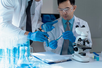 Two scientists wearing lab coats and gloves analyzing test tubes with blue liquid next to a...