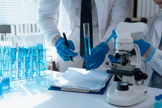 Closeup of scientists analyzing test tube with blue liquid and taking notes on clipboard in a laboratory