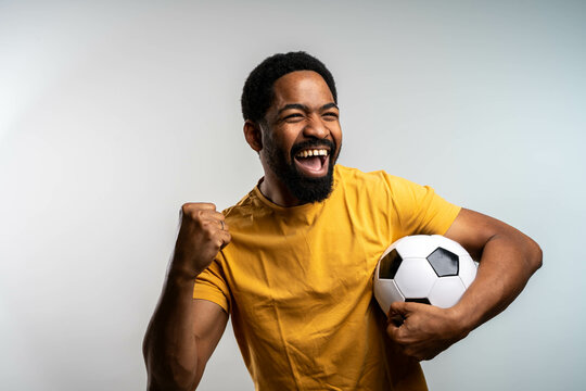 Joyful happy young man in blue shirt posing isolated on grey background. People sincere emotions lifestyle concept. Mock up copy space. Holding soccer ball doing winner gesture screaming.