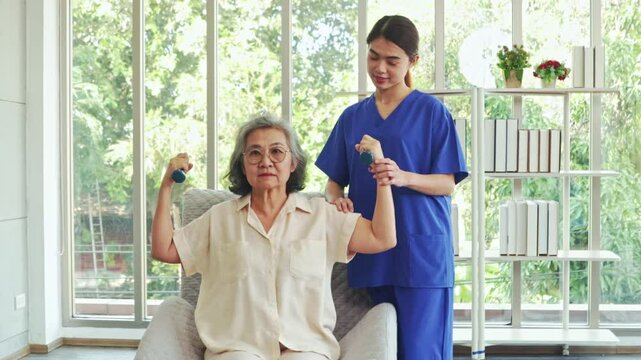 Female physiotherapist is providing nursing care an elderly female patient with muscular dystrophy training her to do dumbbell exercises rehabilitate her muscles and prevent beriberi.