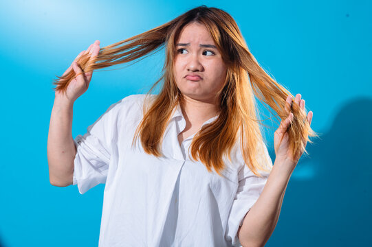Asian woman in a white shirt pulling both sides of her long hair with a sad and frustrated expression, as if showing hair problems or dissatisfaction, against a vibrant blue background