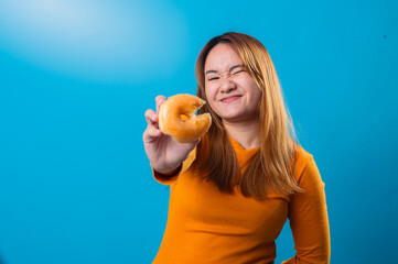Asian woman in an orange shirt holding a bitten glazed donut toward the camera while winking and...