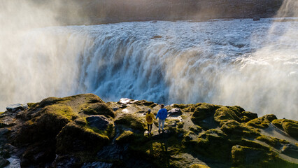Visitors stand on a rocky outcrop, gazing in awe at the powerful Dettifoss waterfall in Iceland. The sun casts a golden light over the cascading water, creating a breathtaking natural spectacle.