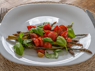 Roasted asparagus topped with juicy cherry tomatoes and basil leaves, served on a clean white plate, fresh and healthy.
