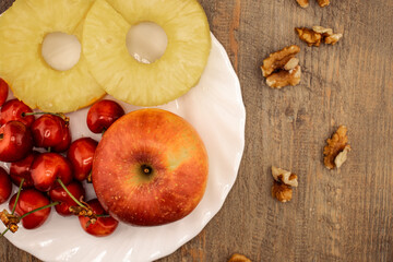 A red apple, pineapple rings, and red cherries are neatly arranged, with scattered walnuts adding a wholesome touch. This image perfectly captures a healthy and delicious snack. 
