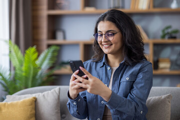 Smiling young Indian woman sitting on sofa at home and using mobile phone