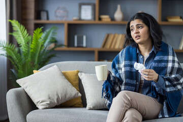 Tired and sick Indian young woman sitting at home on the couch wrapped in a blanket, holding a cup...