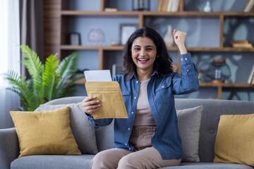 Portrait of a young Indian woman sitting on the sofa at home. Holding an open envelope with a letter in her hand and rejoicing in success, raising her hand up