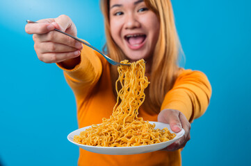 Asian woman in orange sweater smiling and offering a forkful of instant noodles towards the camera...