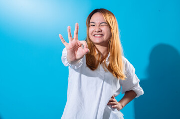 Asian woman with long straight hair in a white shirt smiles brightly while making an OK hand...