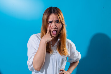 Asian woman in a white shirt pulling her lower eyelid down with one finger and sticking out her tongue in a silly and exaggerated expression, standing against a solid blue background