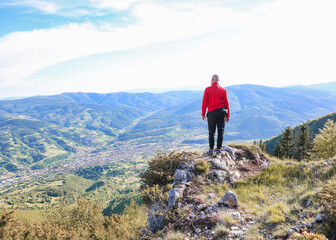 Obraz premium Man in red jacket standing on a mountain peak, looking at the panoramic valley below — concept of freedom, achievement, perspective, and connection with nature.