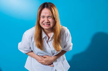 Asian woman in white shirt holding her stomach in pain with a tense facial expression, eyes closed and teeth clenched, expressing abdominal discomfort in front of a solid blue background
